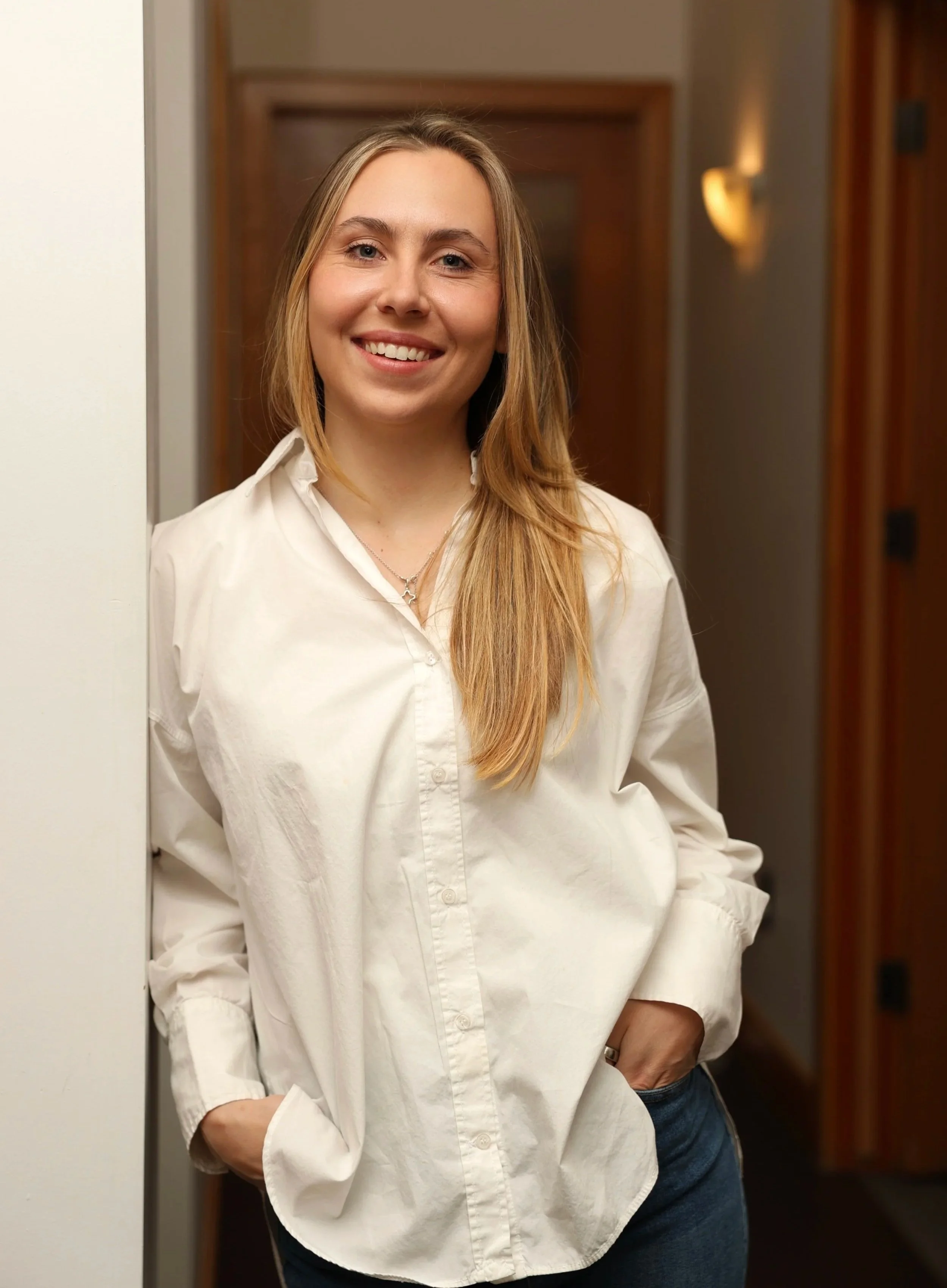 A smiling woman with long blonde hair, wearing a white button-up shirt and blue jeans, standing in a hallway with wooden doors and wall-mounted lights.