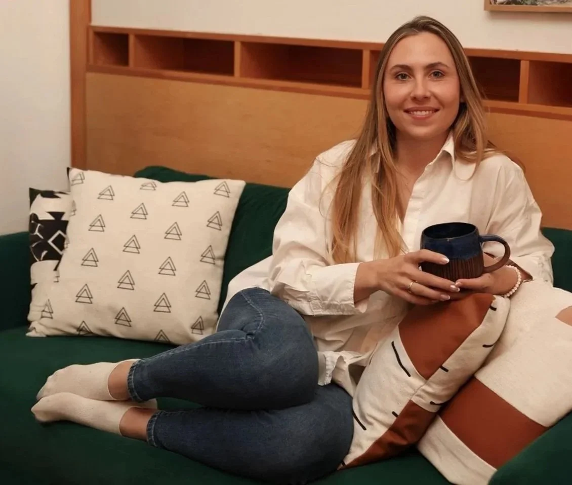 A woman sitting on a green sofa, smiling, holding a dark blue mug, wearing a white shirt and jeans, with pillows behind her, in a room with wood-paneled wall.