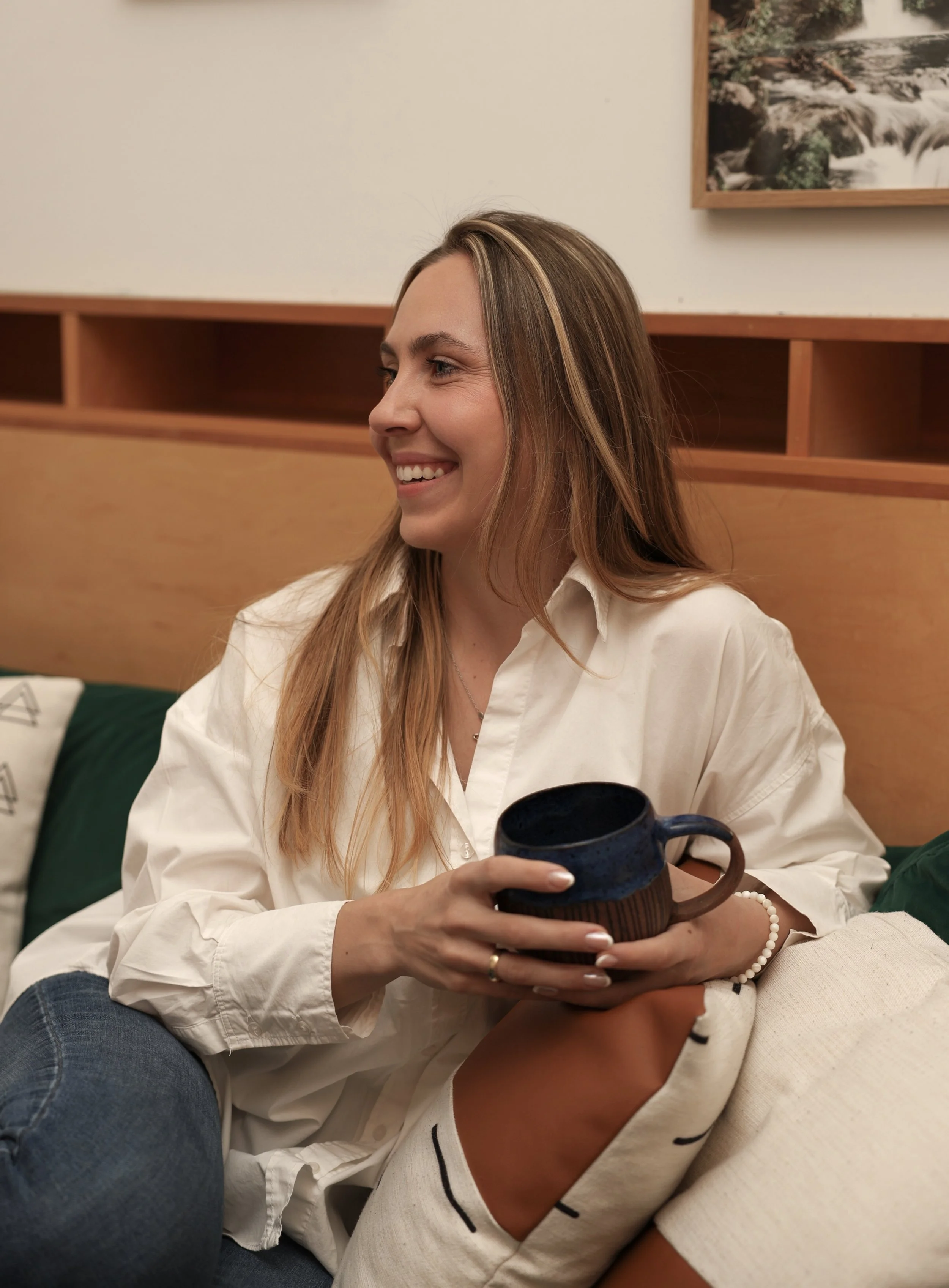 A woman with long, light brown hair smiling while sitting on a couch, holding a dark blue coffee mug.