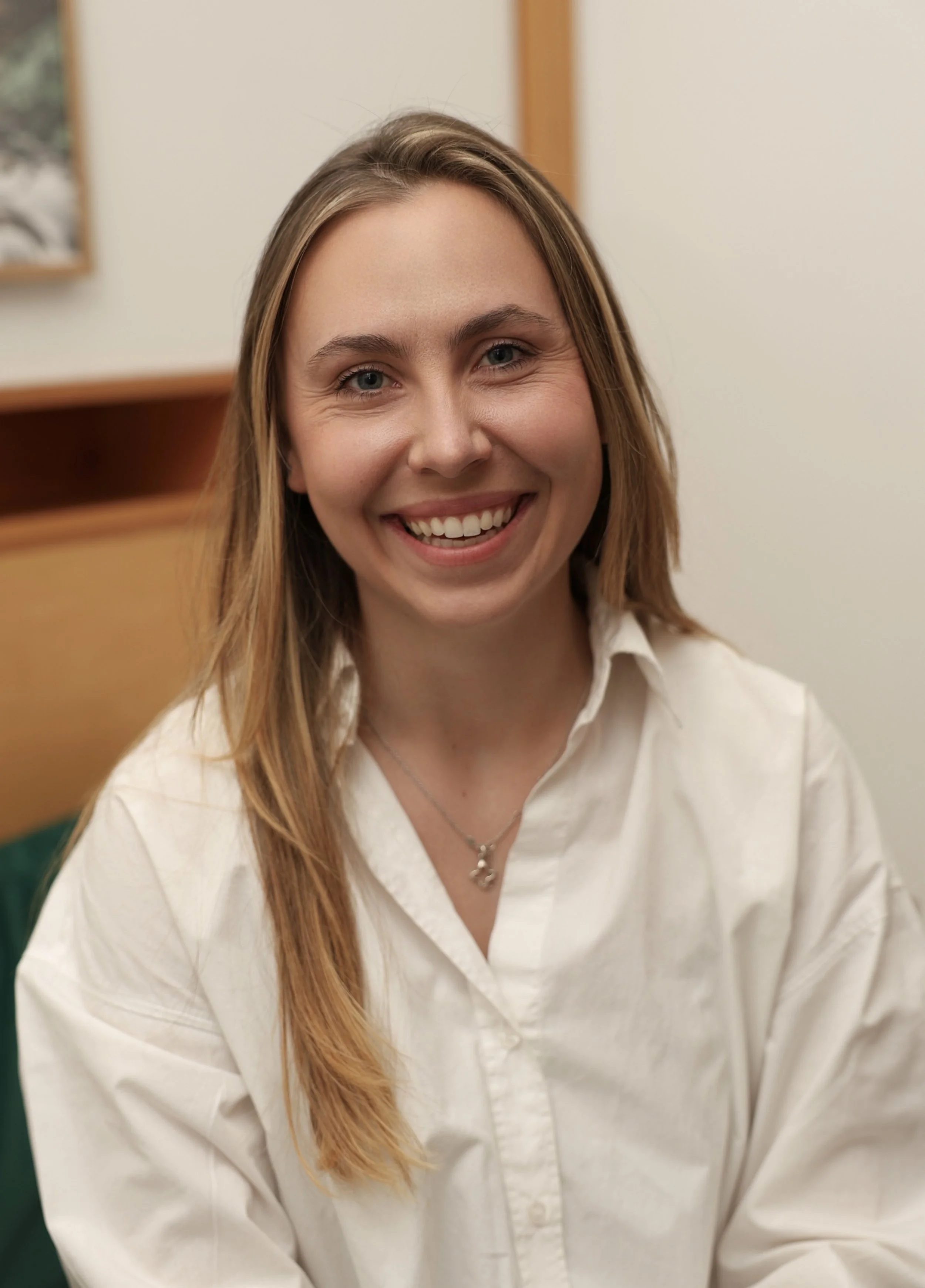 A young woman with long, light brown hair smiling at the camera, wearing a white button-up shirt and a silver necklace, seated indoors.