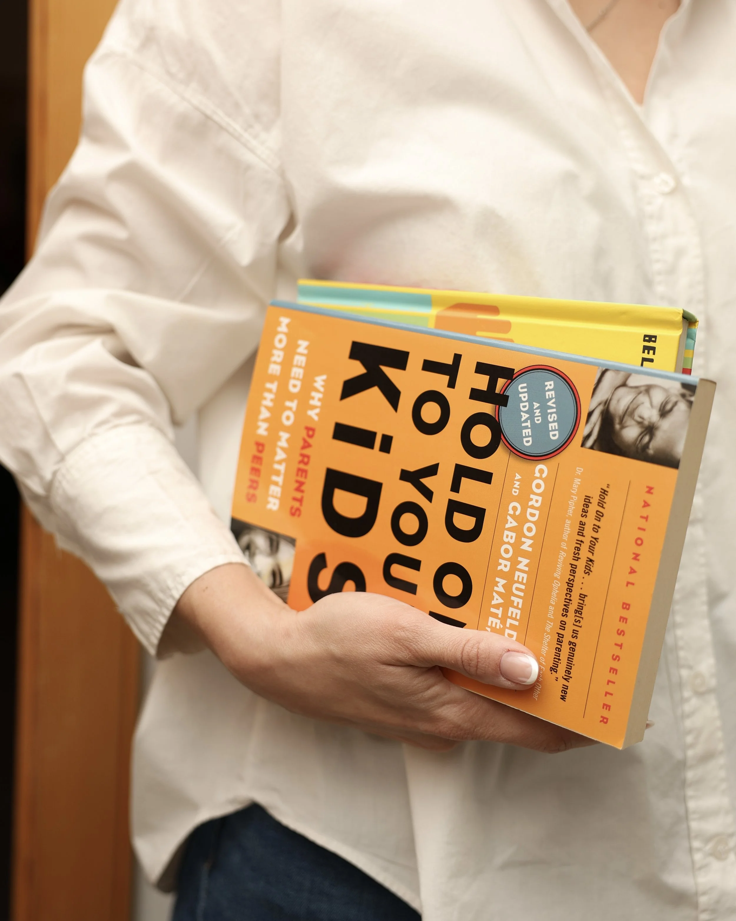 A person wearing a white shirt holding a colorful book titled 'Hold On To Your Kids' by Gordon Neufeld and Gabor Mate, with visible text indicating it is a revised and updated edition.