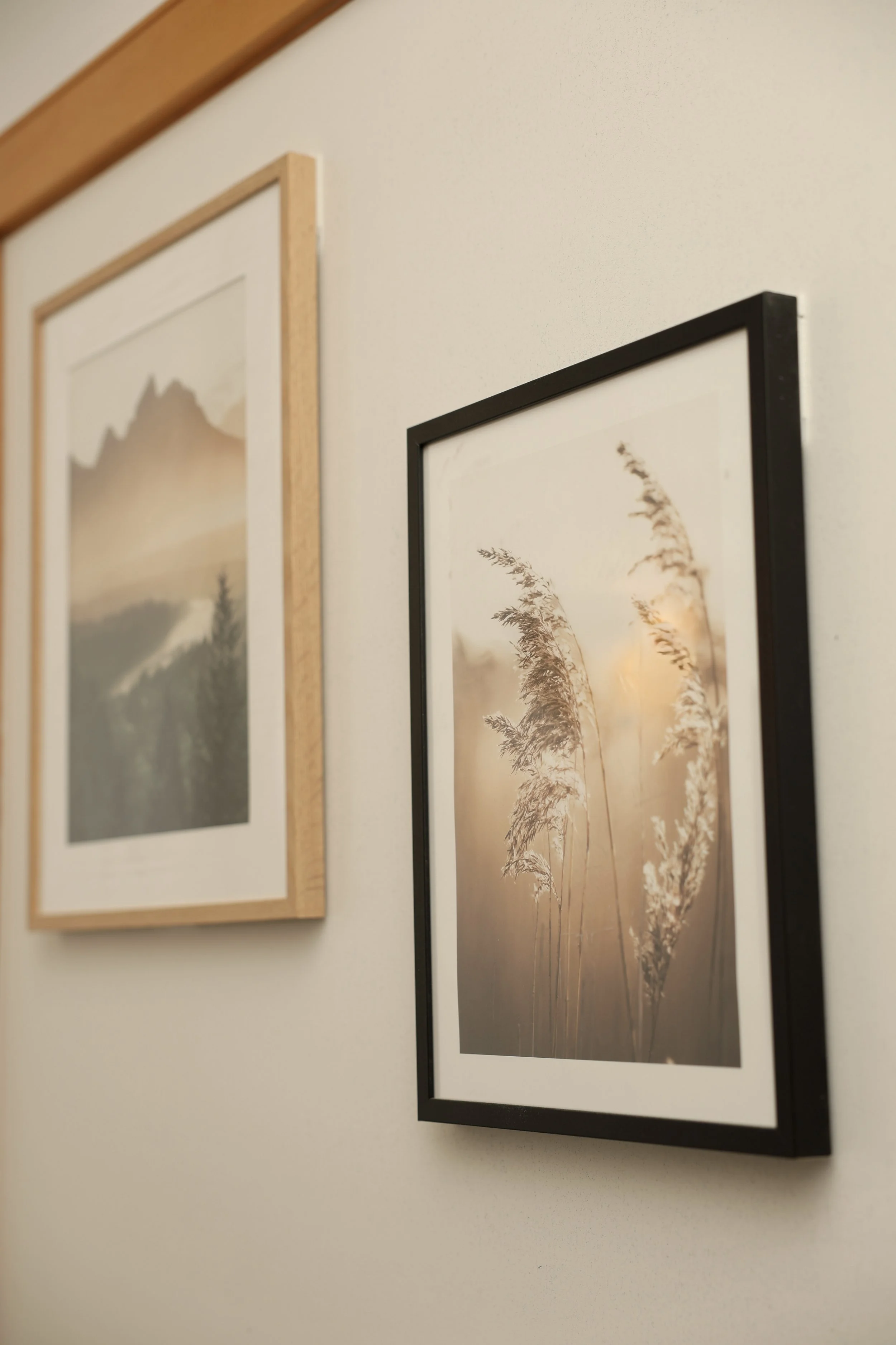 Two framed landscape photographs hung on a white wall. The photo on the right features tall grasses in a soft, warm light. The photo on the left depicts a mountain scene.