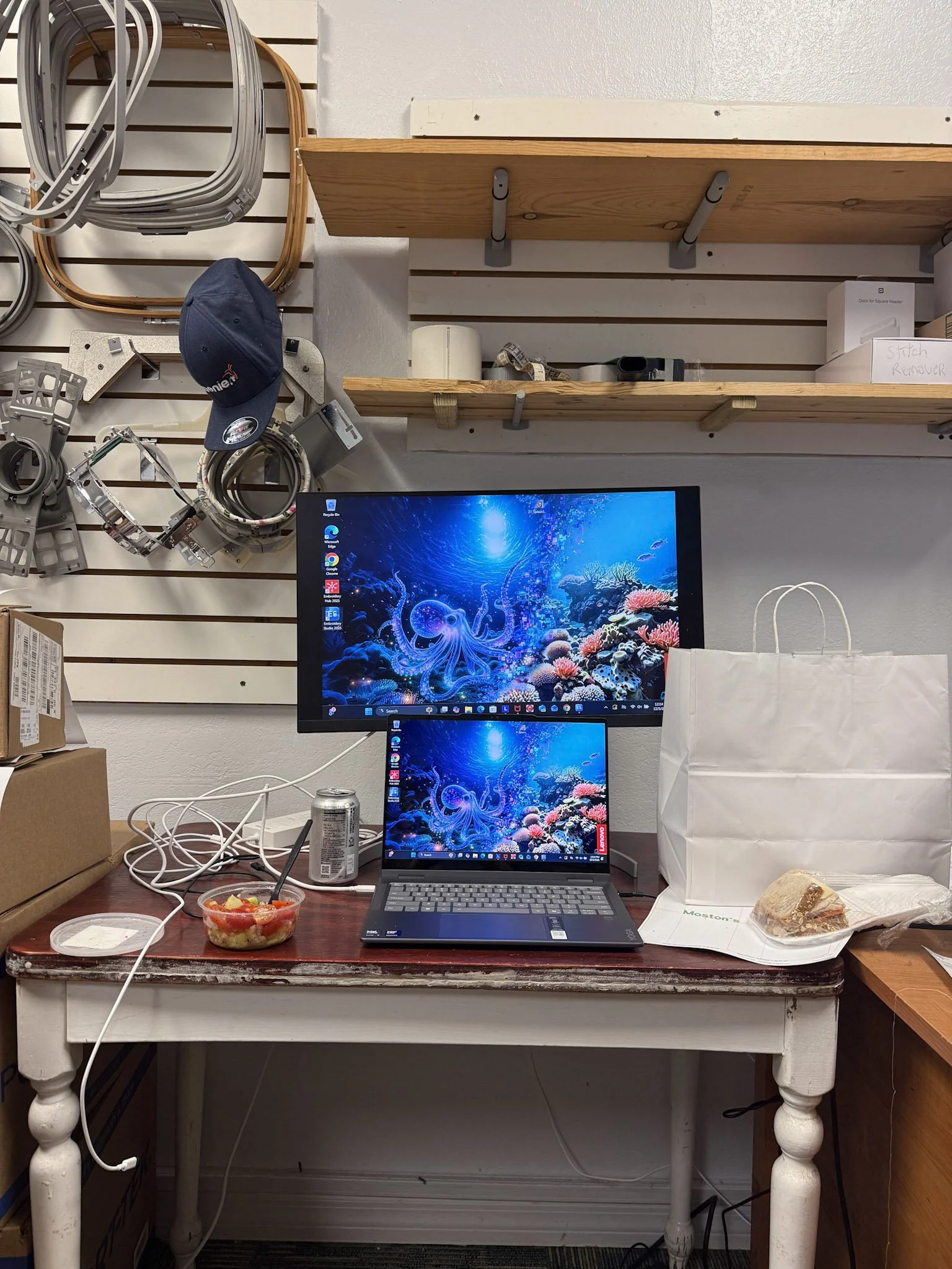 A cluttered workspace with a laptop and dual monitors displaying an underwater scene, a bowl of food, a soda can, and a paper bag on a distressed white table. Behind, there is a wall with wooden shelves and various cords and hardware hanging on a pegboard.