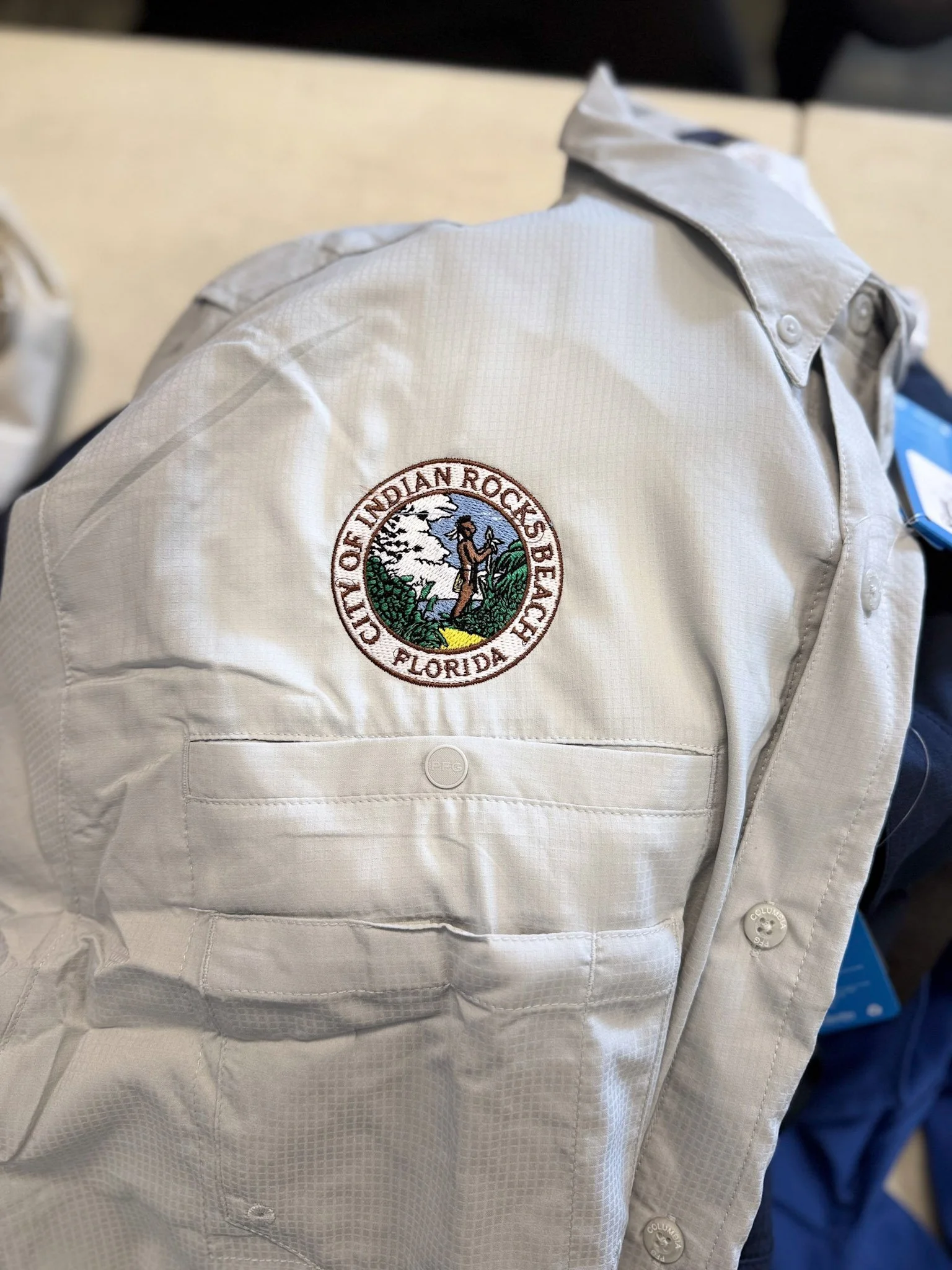 A beige uniform shirt with a circularpatch that says 'City of Indian Rocks Beach, Florida' and depicts a person standing in nature with trees and clouds.