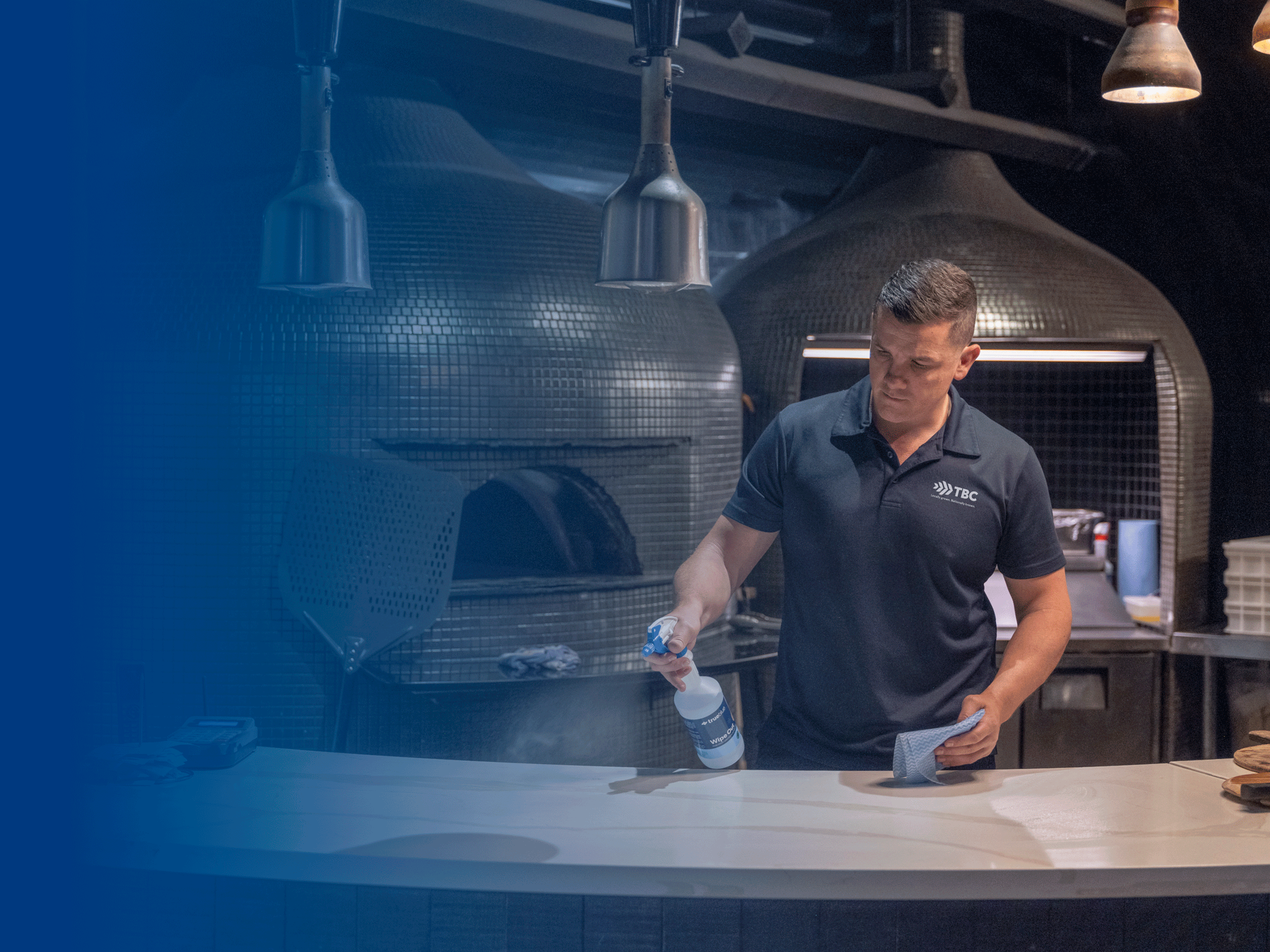 A man cleaning a countertop in a pizza restaurant kitchen with large ovens in the background.