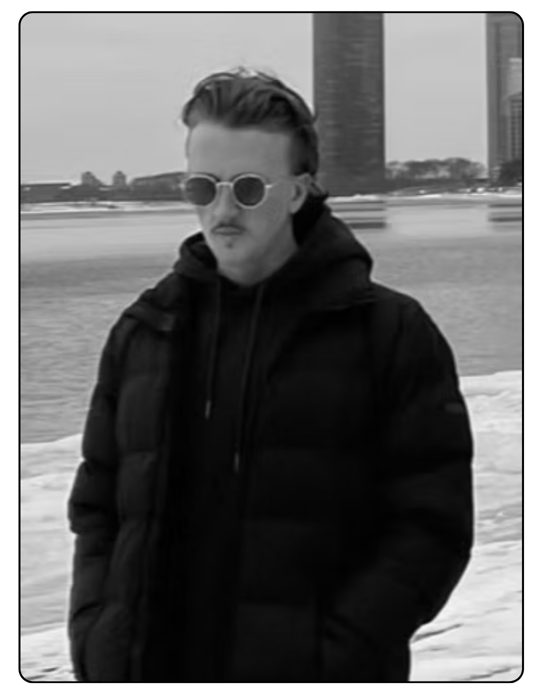A man standing outdoors in winter, wearing sunglasses, a dark puffy jacket, and a hoodie, with snow on the ground and city buildings in the background.