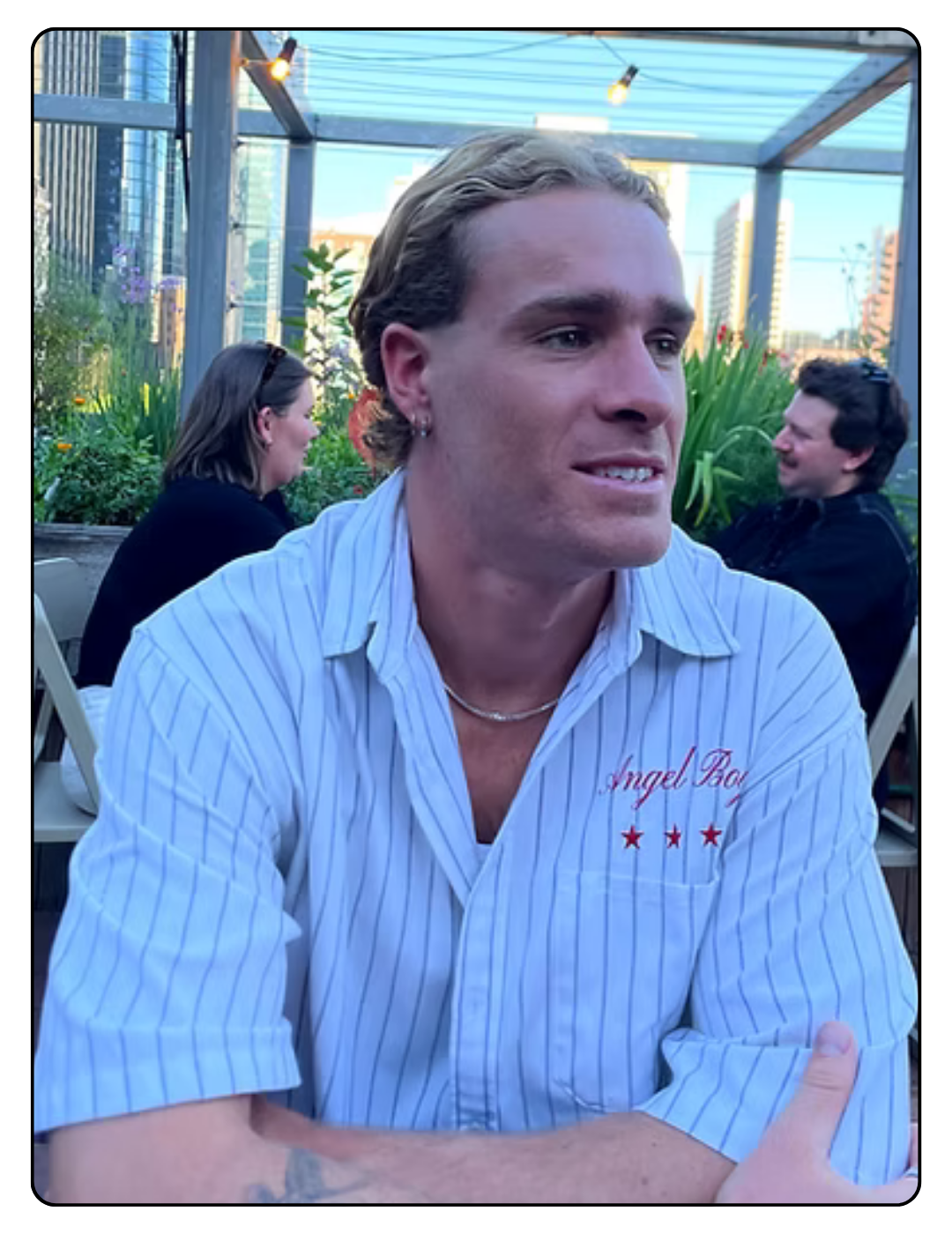 A young man with long blonde hair sitting at an outdoor patio, smiling slightly, wearing a blue striped shirt with 'Angel Bay' embroidered on it, and a pearl necklace. There are two women in the background, one with dark hair and sunglasses on her head, and the other with short dark hair, both seated at the patio among plants and city buildings.