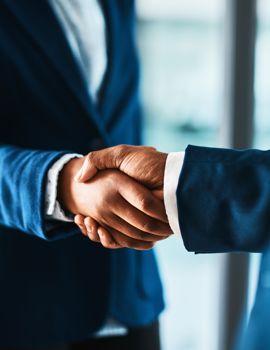 Two individuals shaking hands, both wearing business suits, in a professional setting.