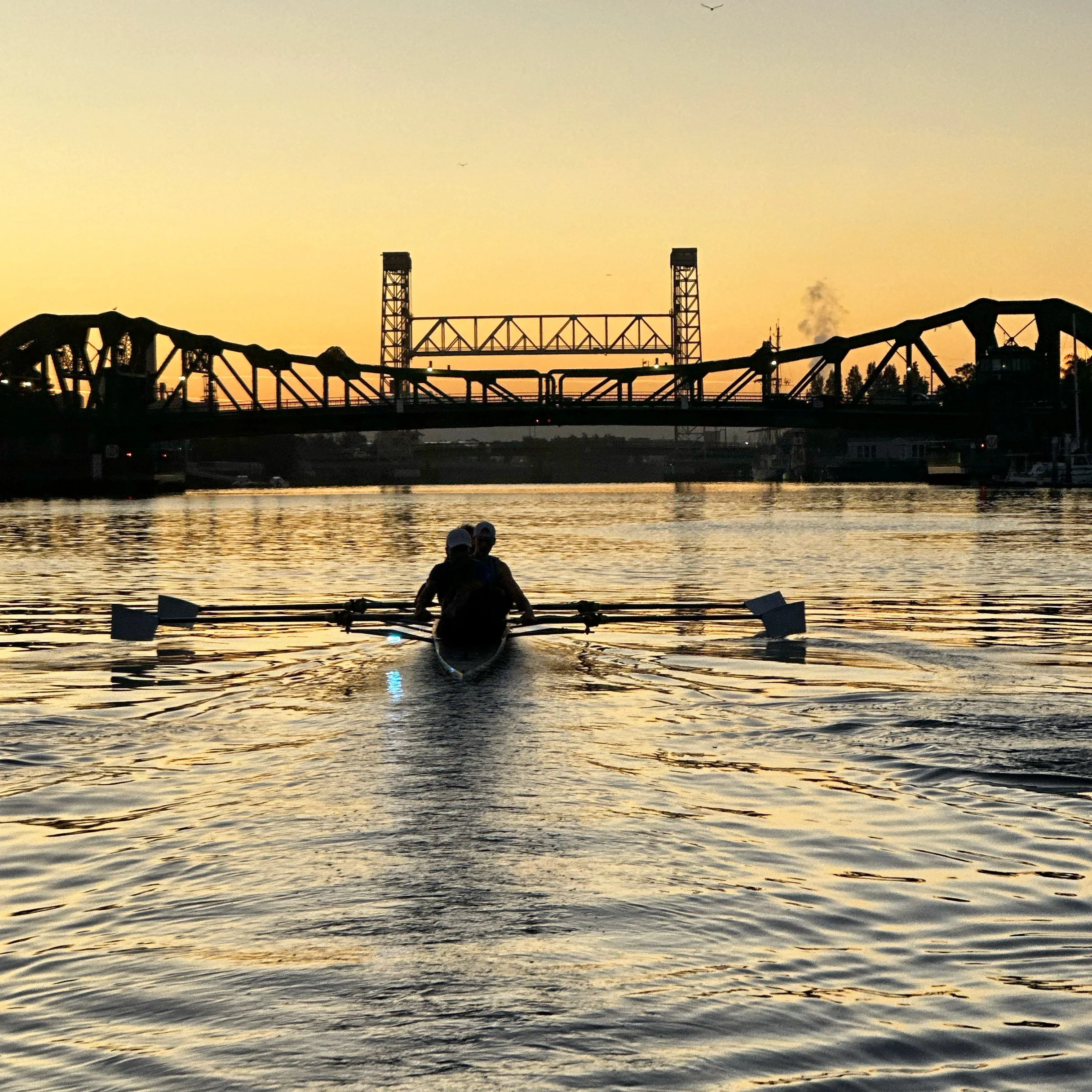 Two rowers paddle a boat on a river during sunset, with a bridge in the background and two silhouetted skydivers in the sky.