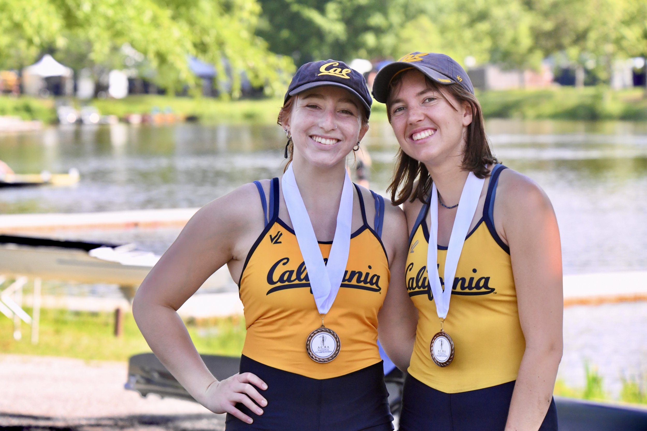 Two women wearing matching yellow and blue California rowing uniforms and caps with medals around their necks, standing outdoors by a water body with trees in the background, smiling and posing for the photo.