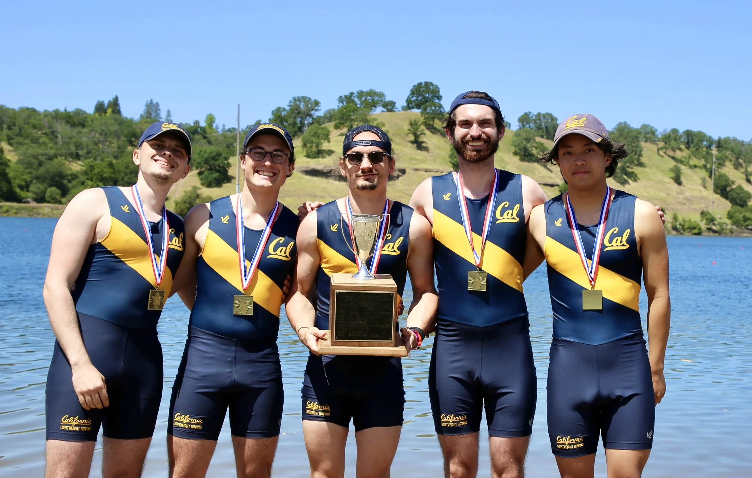 A group of five young men in California rowing team uniforms happily standing by a lake with trees and hills in the background. They are holding a trophy and wearing medals.