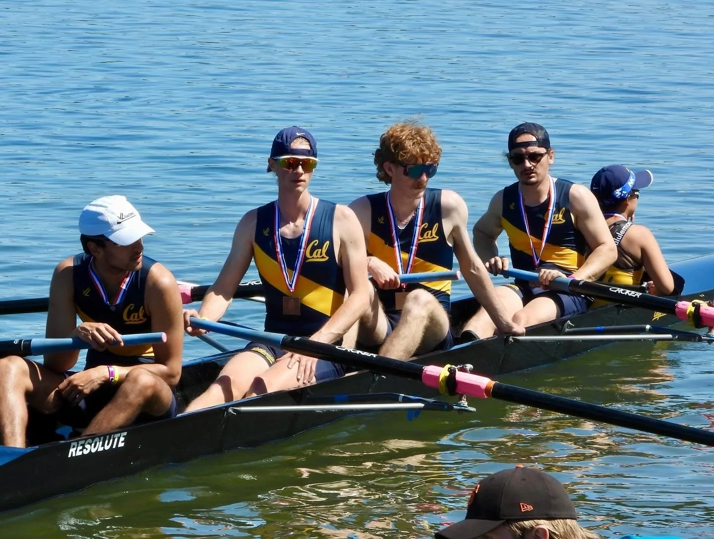 A team of five rowers in a black and yellow boat on a lake, wearing matching Cal team uniforms and medals.