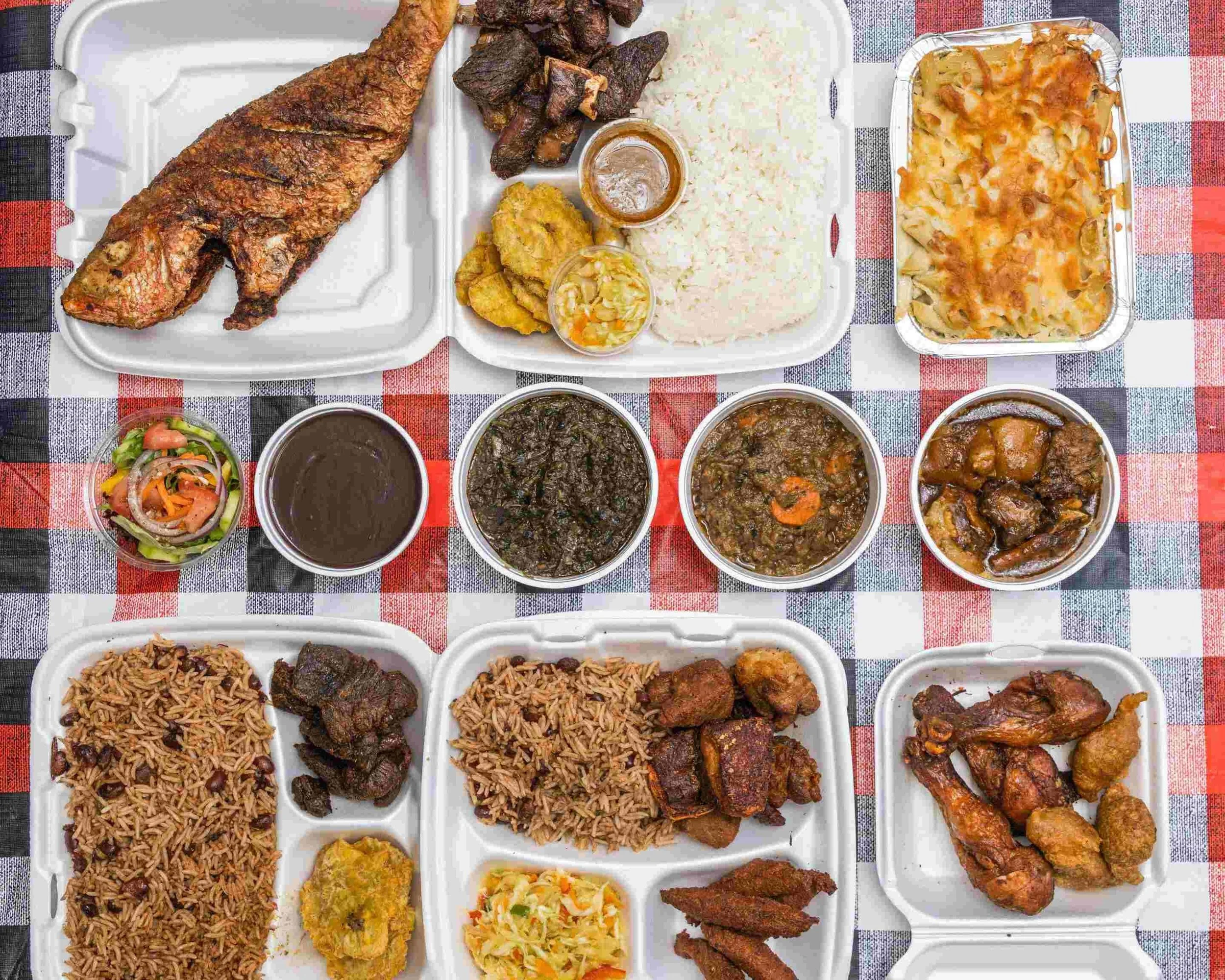 A spread of Haitian food on a checkered tablecloth, including fried fish, chicken, griot, rice, beans, plantains, salad, and soups.