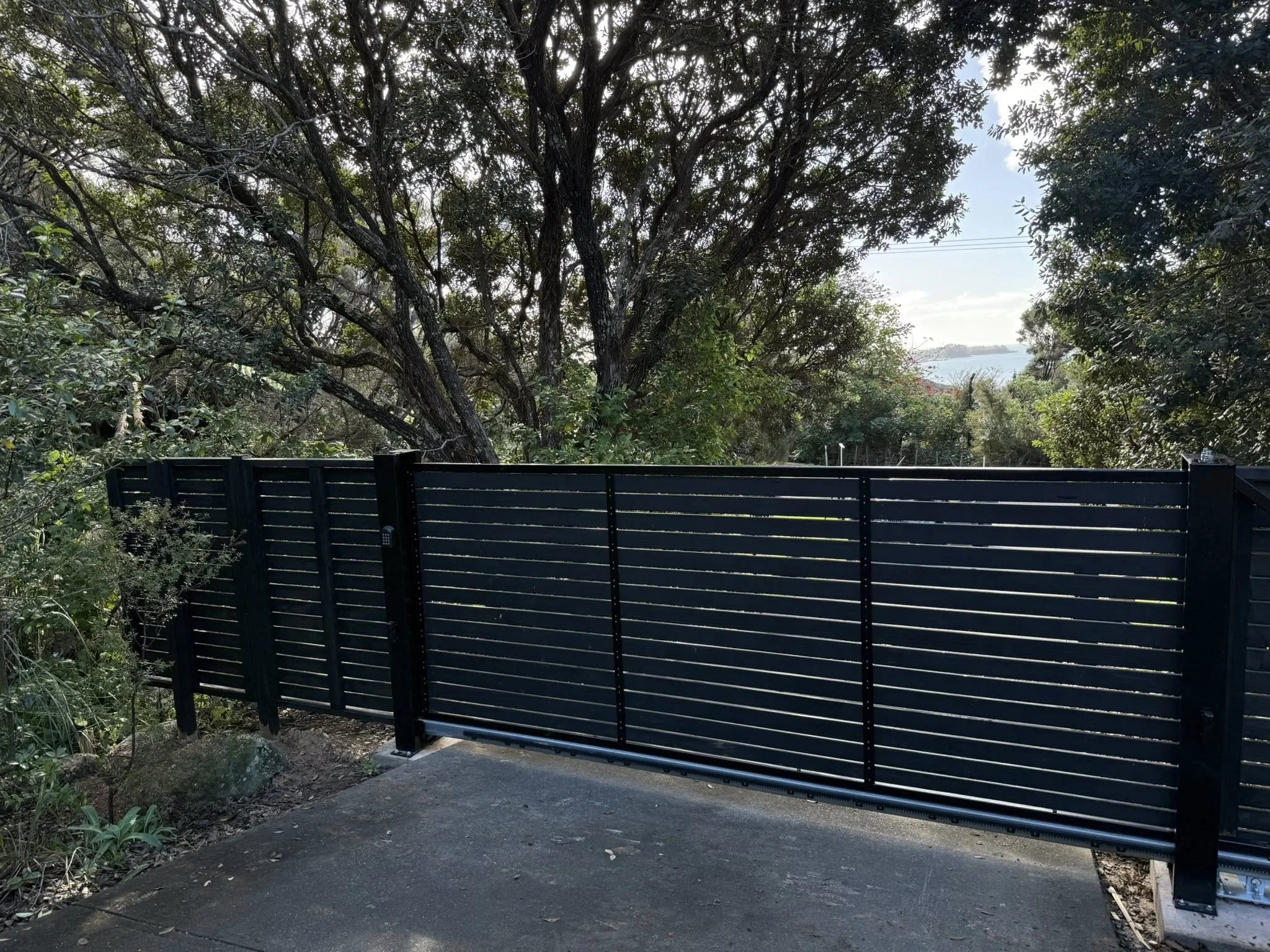 Black metal gate with horizontal slats in a natural setting with trees and bushes, and a view of water and sky in the background.