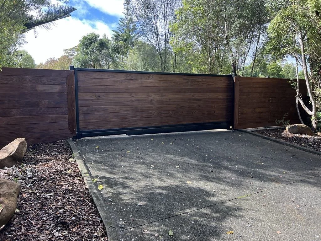 Wooden driveway gate with metal frame in a suburban yard, surrounded by trees and rocks, with a concrete pathway leading to the gate.
