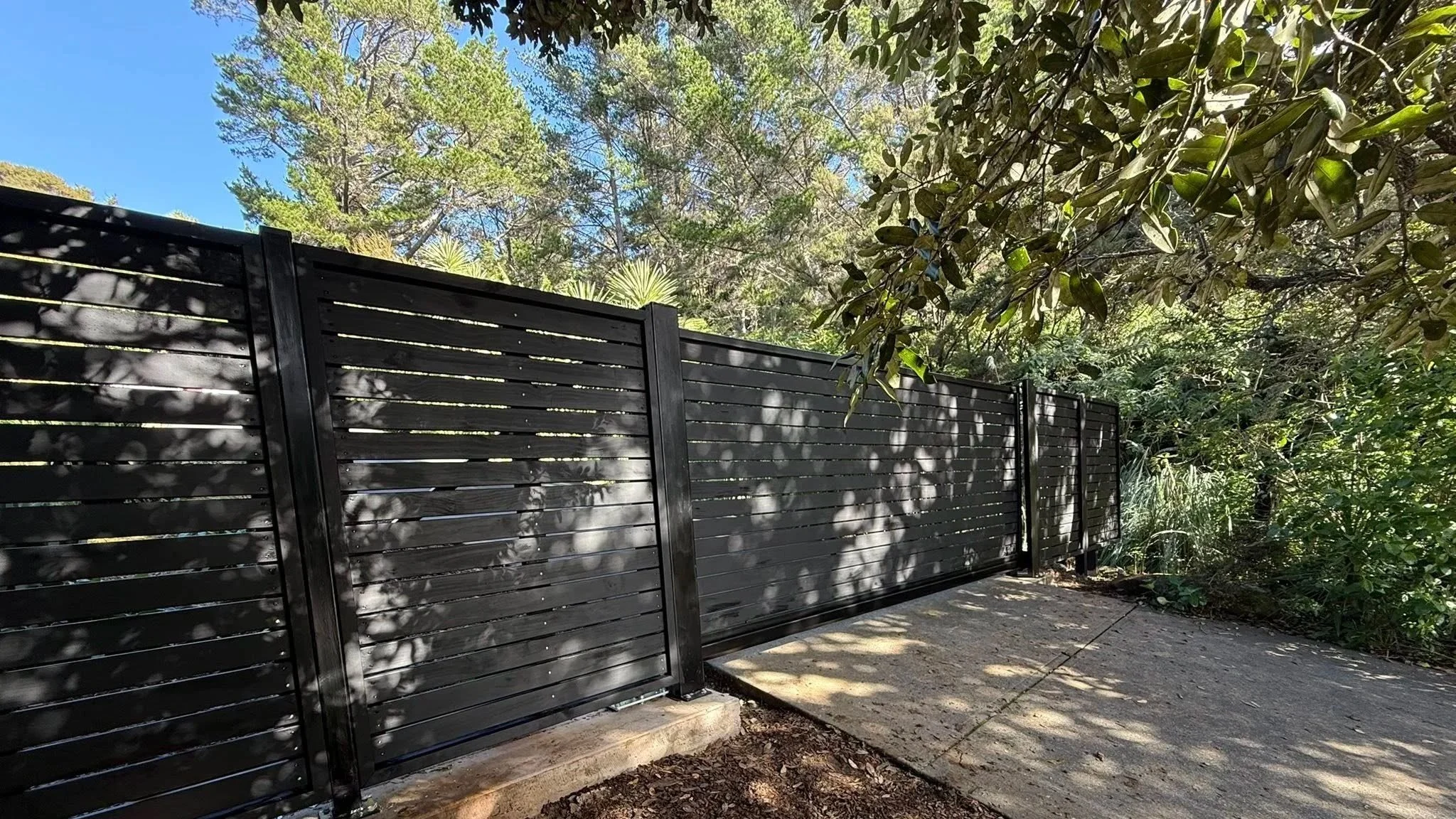 A black metal gate with horizontal slats, surrounded by green trees and bushes, with a concrete pathway in front and a clear blue sky above.