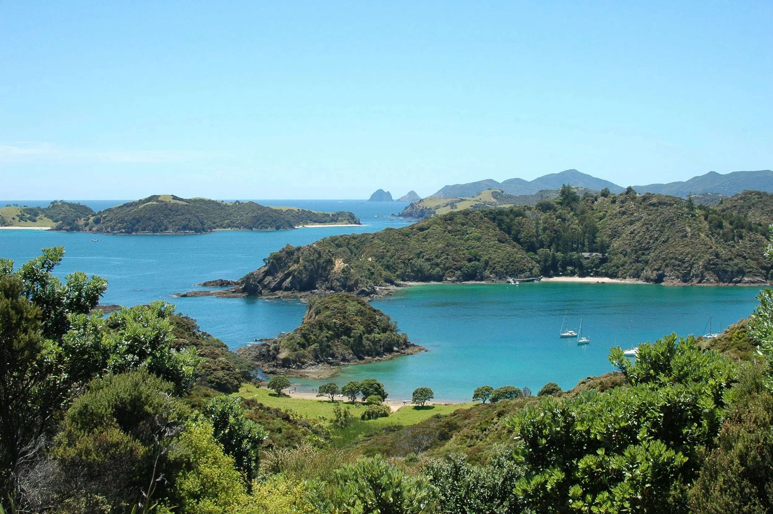 Scenic view of a bay with turquoise waters, surrounded by green hills and islands, with sailboats anchored near the shore and a distant mountainous coastline under a clear blue sky.