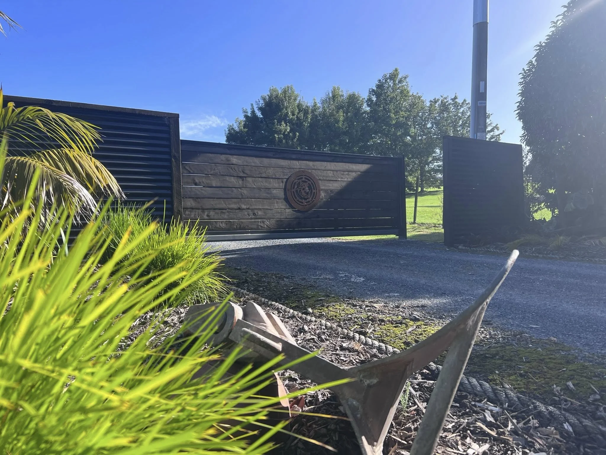A closed black wooden gate with a decorative circular emblem at the center, flanked by two black wooden fences. Green bushes and plants are in the foreground, and a grassy area with trees is visible in the background under a clear blue sky.
