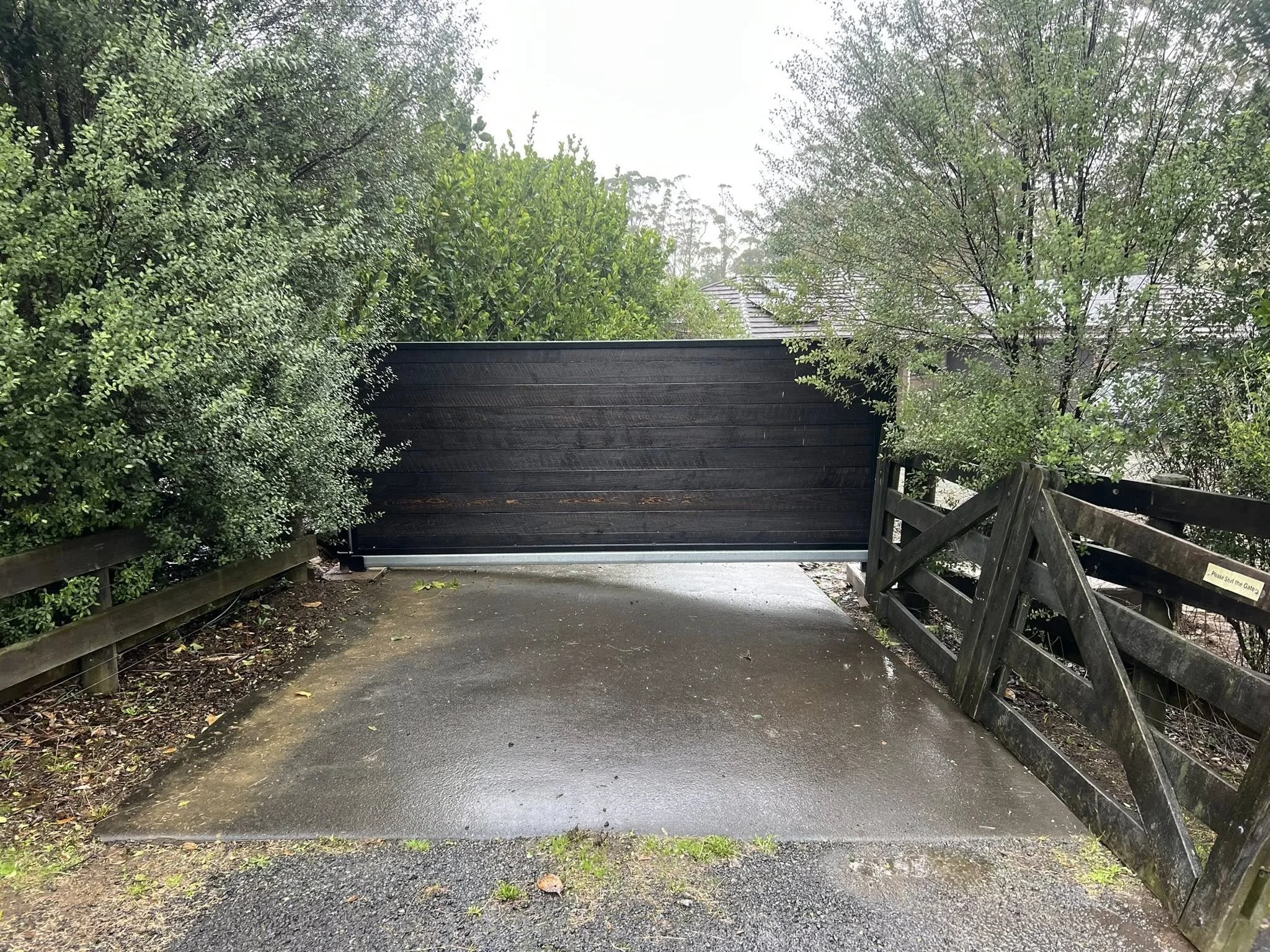 A black metal gate blocking a driveway, with wooden fences on either side, surrounded by green bushes and trees on a rainy day.
