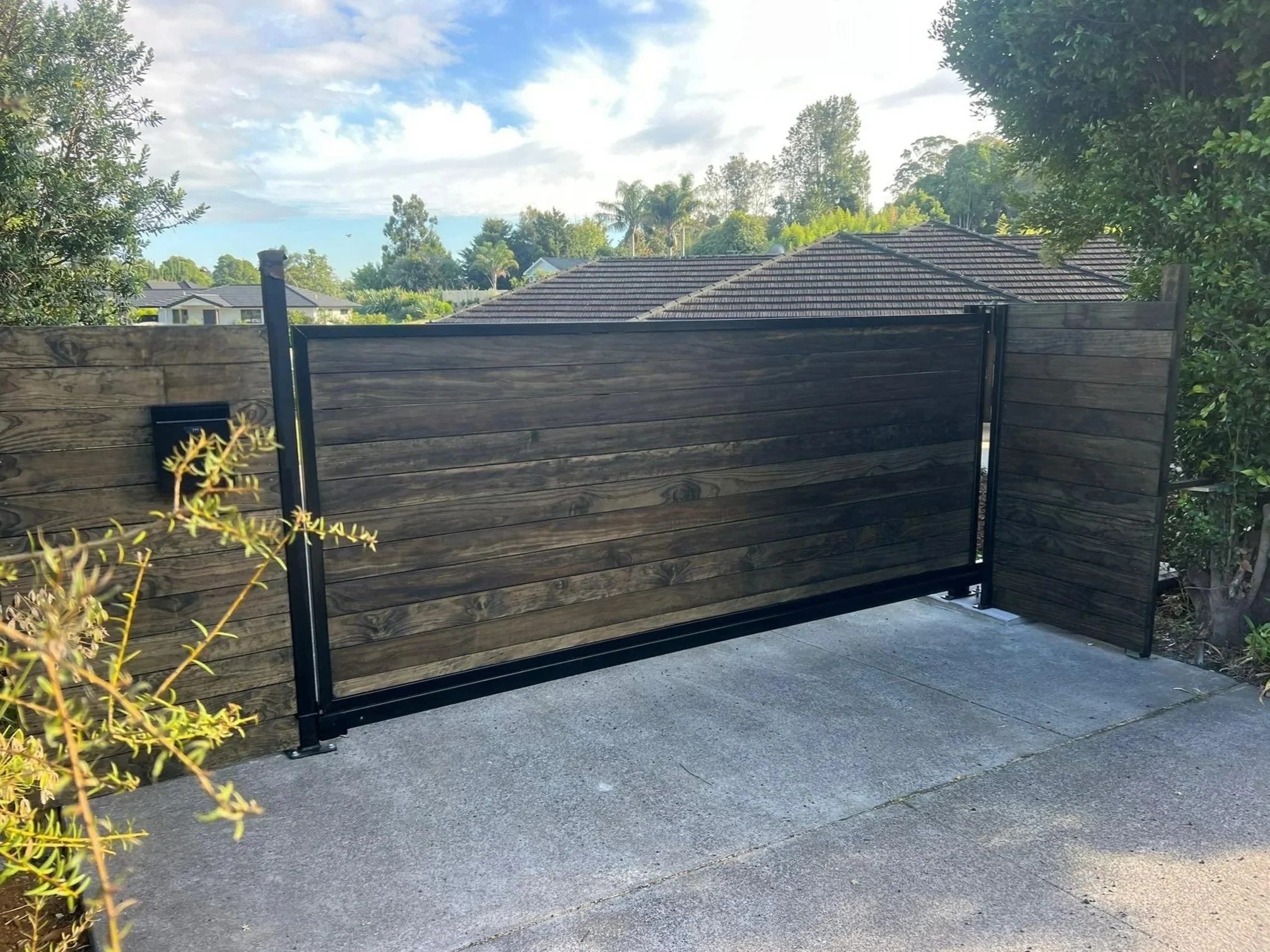 A black metal gate with wooden panels, attached to a wooden fence on one side, on a concrete driveway with greenery and trees in the background.
