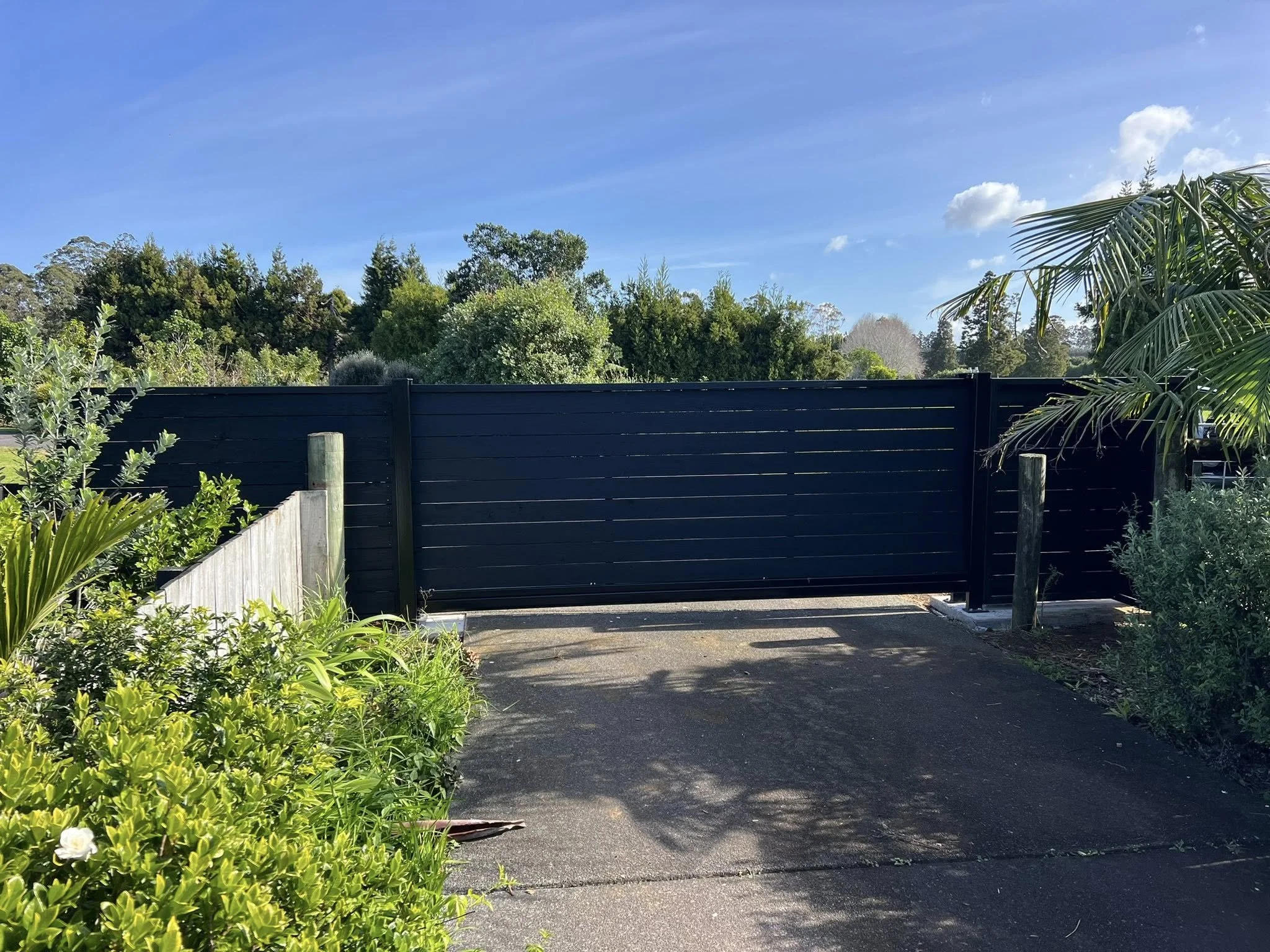 A black gate with a driveway in front, surrounded by green bushes and trees, under a clear blue sky with some white clouds.