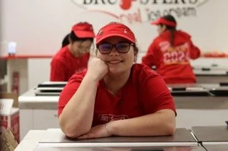 Young woman sitting at a table in a Burger King restaurant, wearing glasses and a red cap, resting her head on her hand, smiling, with employees in red uniforms in the background.