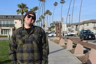 Man in sunglasses and plaid shirt standing on sidewalk with palm trees and apartment buildings in the background