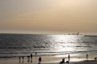 People walking along the beach at sunset, with a ship in the distance on calm water.