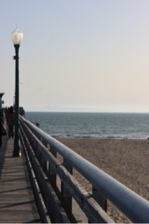 View of a wooden pier with white railing and lampposts, extending over a beach toward the ocean with a hazy sky.