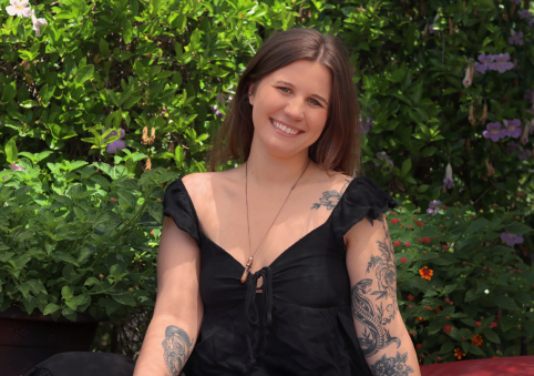 Smiling young woman with tattoos on arms and chest, wearing a black top, standing outdoors against lush green foliage with purple and orange flowers.