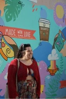 Woman with curly hair wearing a red sweater and patterned skirt smiling in front of a colorful mural with coffee cup and plant illustrations.