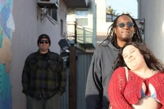 Two men and a woman walking in an alleyway, with the woman leaning on the man on the right and all three wearing casual clothing and sunglasses.