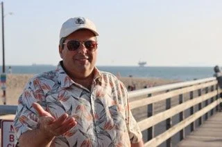Man wearing sunglasses, a cap, and a tropical shirt, standing on a boardwalk near the beach, making a gesture with his hand.
