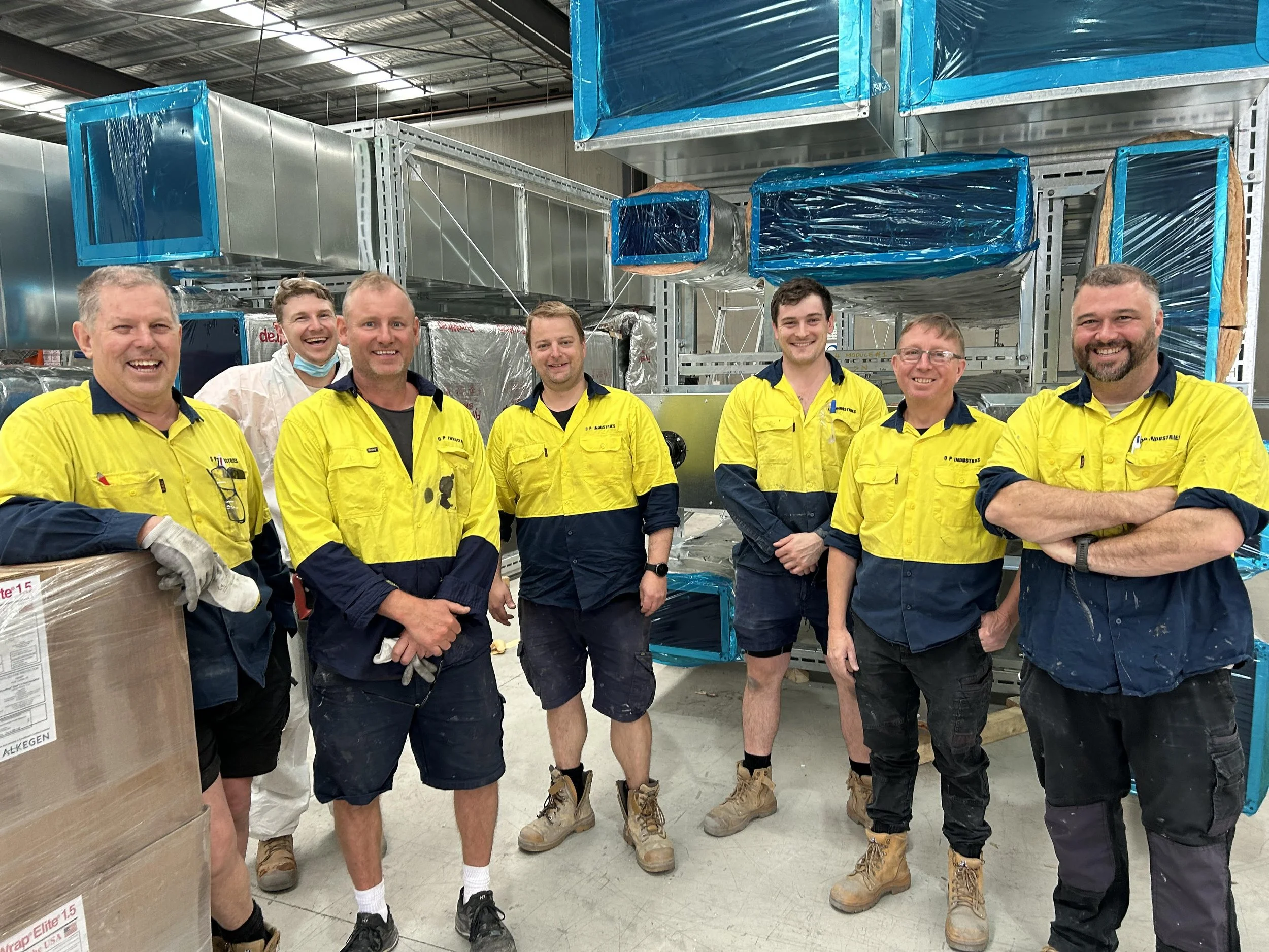 A group of seven workers in yellow and navy uniforms standing inside a warehouse surrounded by equipment and blue-wrapped materials, smiling for the camera.