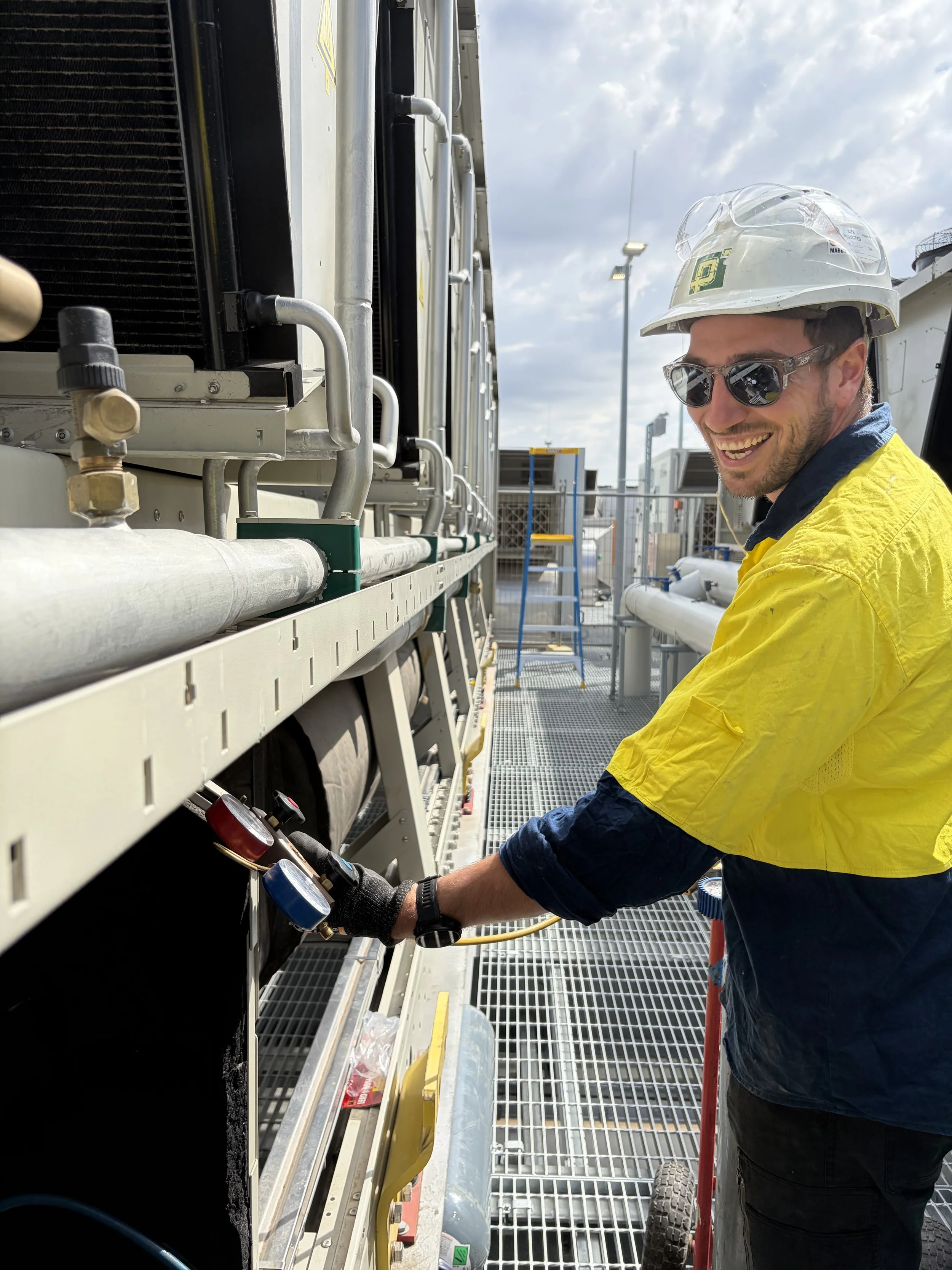 A worker in safety gear, including a hard hat and sunglasses, is checking gauges on industrial equipment outdoors with a cloudy sky in the background.