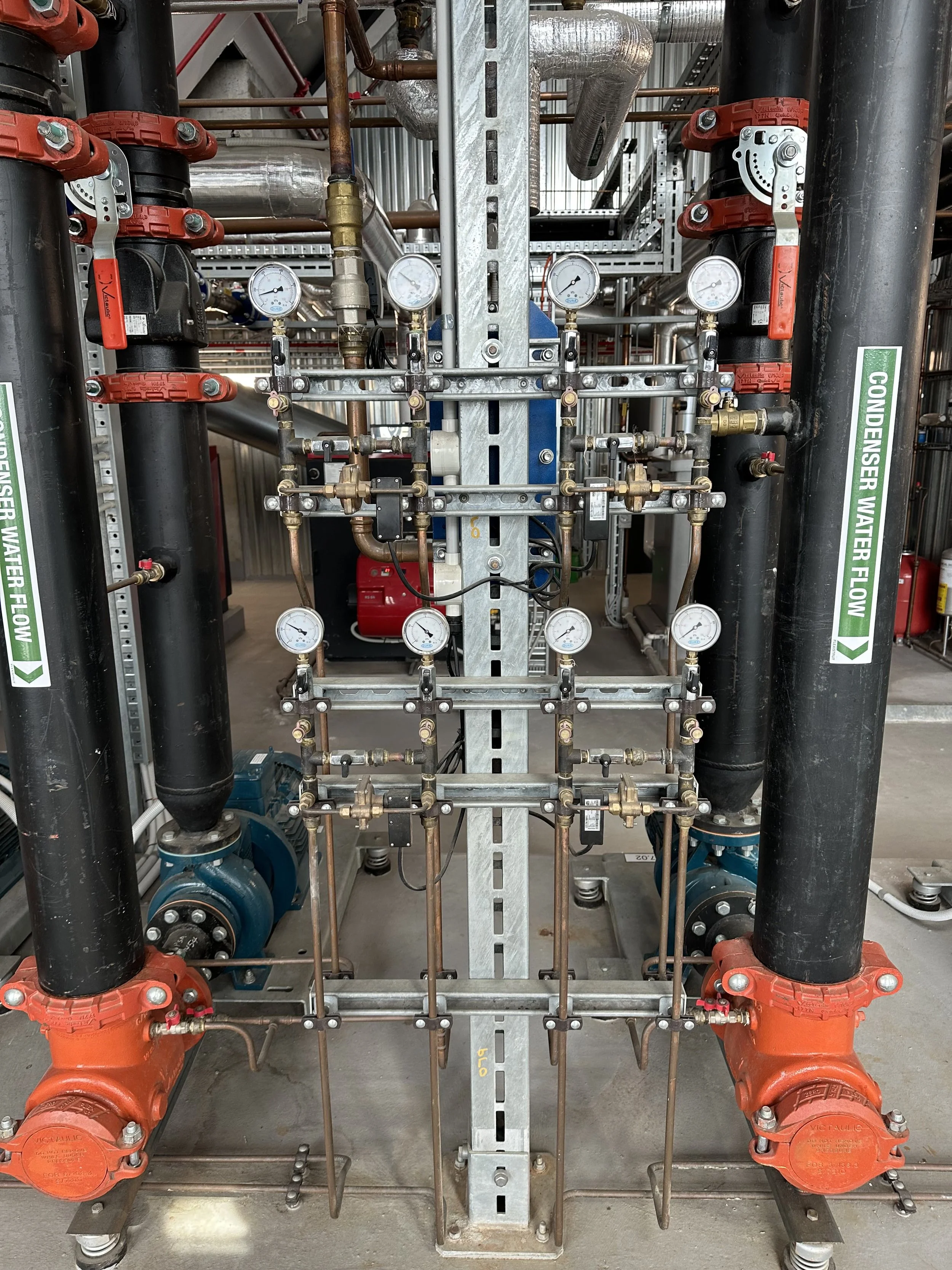 Industrial pipes and gauges for water flow control, labeled 'Condenser Water Flow', with two large black pipes, multiple valves, and pressure gauges installed on a metal frame.