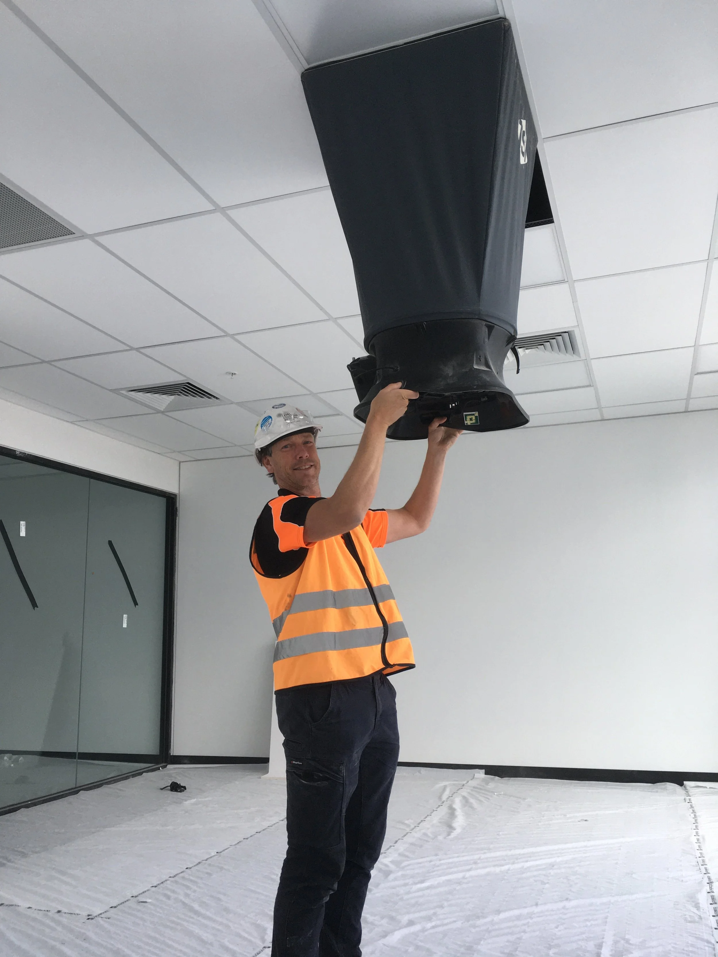 Construction worker in safety vest and hard hat installing or inspecting a large speaker in an indoor office space.