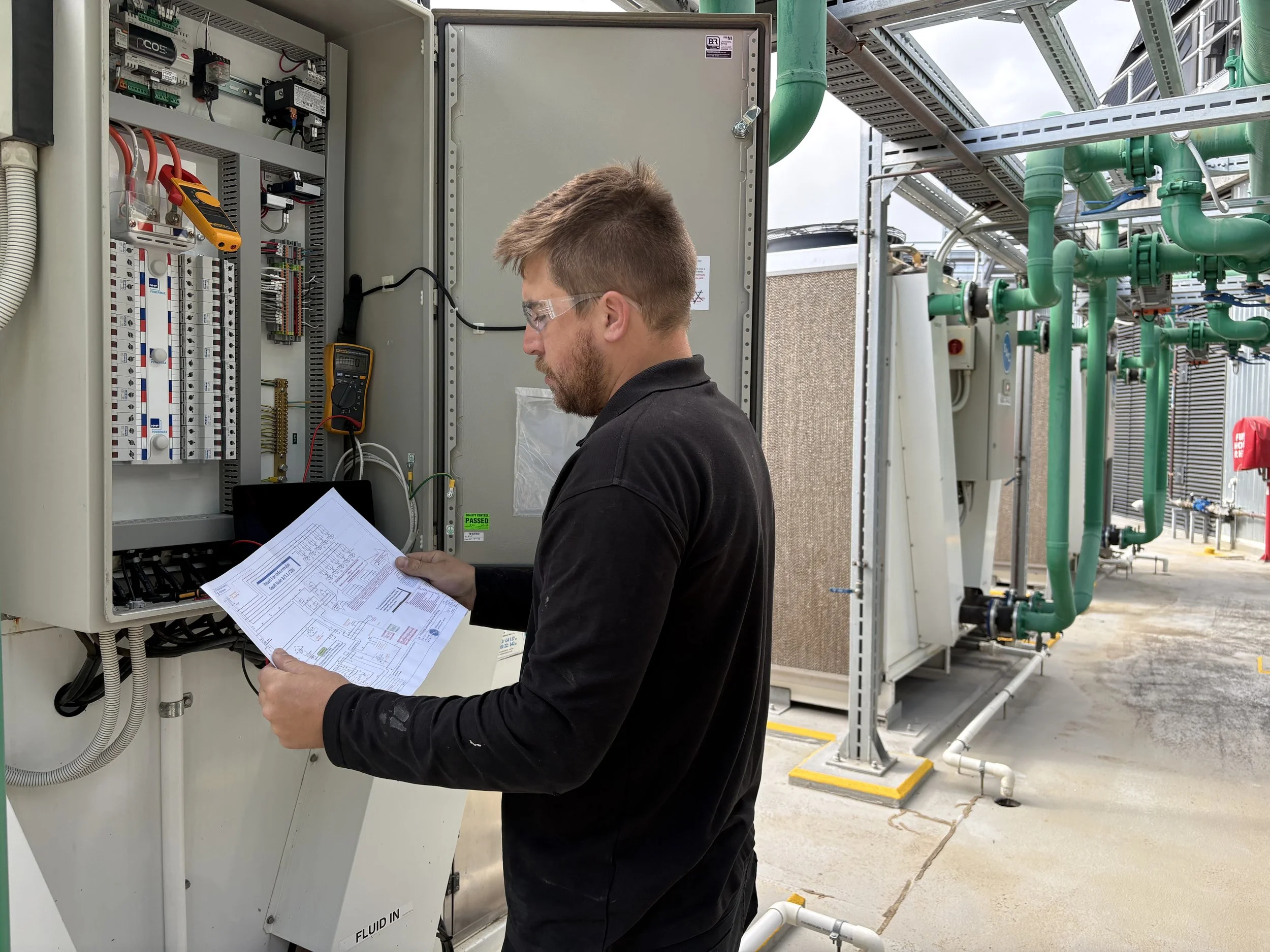 Electrician inspecting an electrical control panel with printed schematics at an industrial outdoor site with green piping and equipment.
