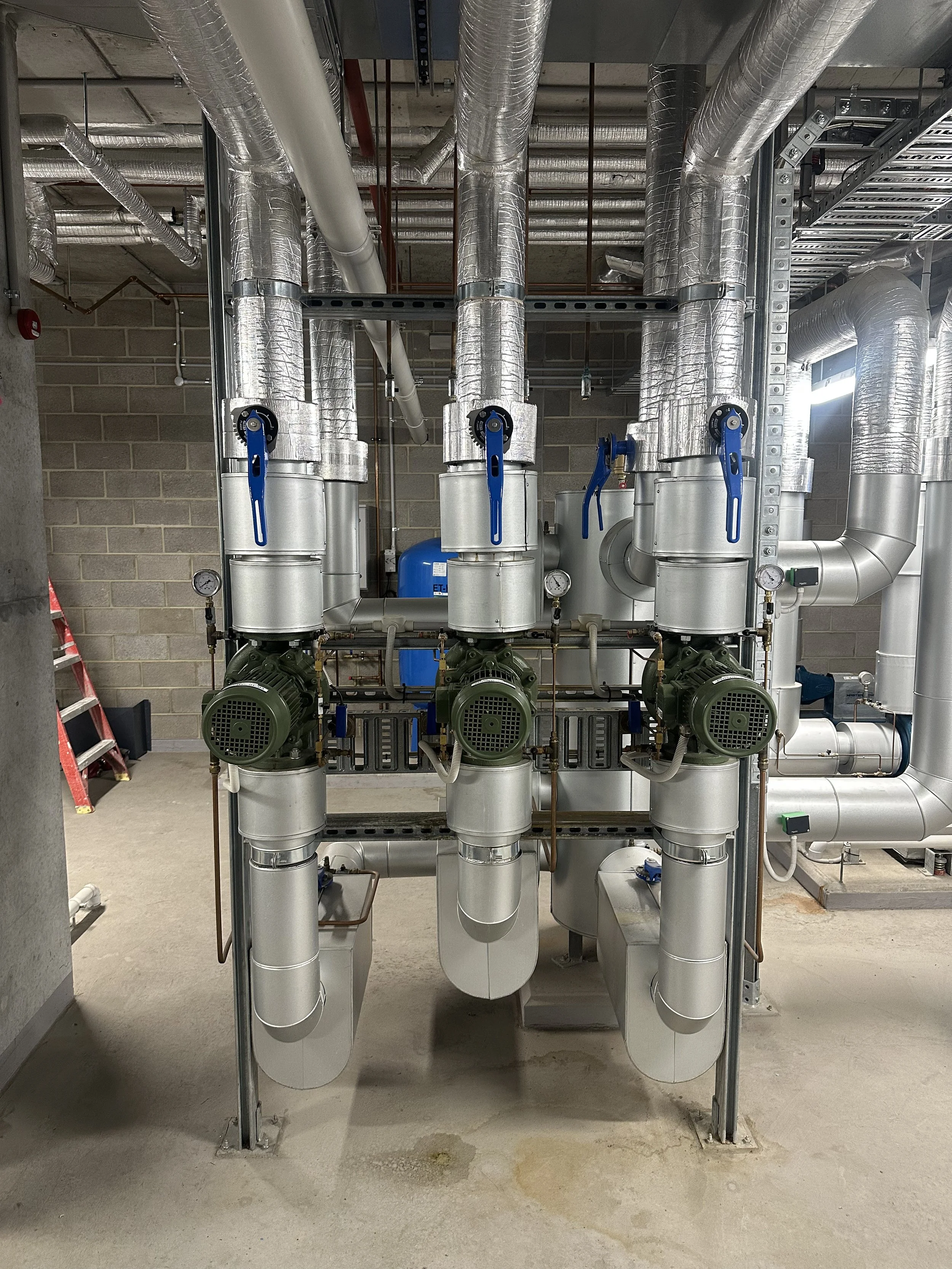 Industrial HVAC or mechanical piping system with large insulated pipes, pumps, gauges, and blue valve handles in a building utility room.