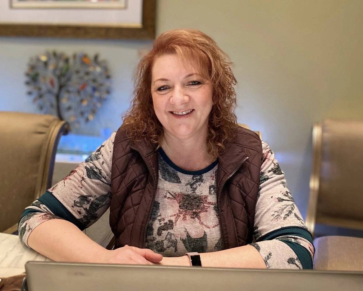 A woman with curly reddish hair smiling at the camera, sitting at a table with a laptop in front of her. She is wearing a floral shirt and a quilted vest, in a room with beige and green walls and a decorative metal wall art piece in the background.