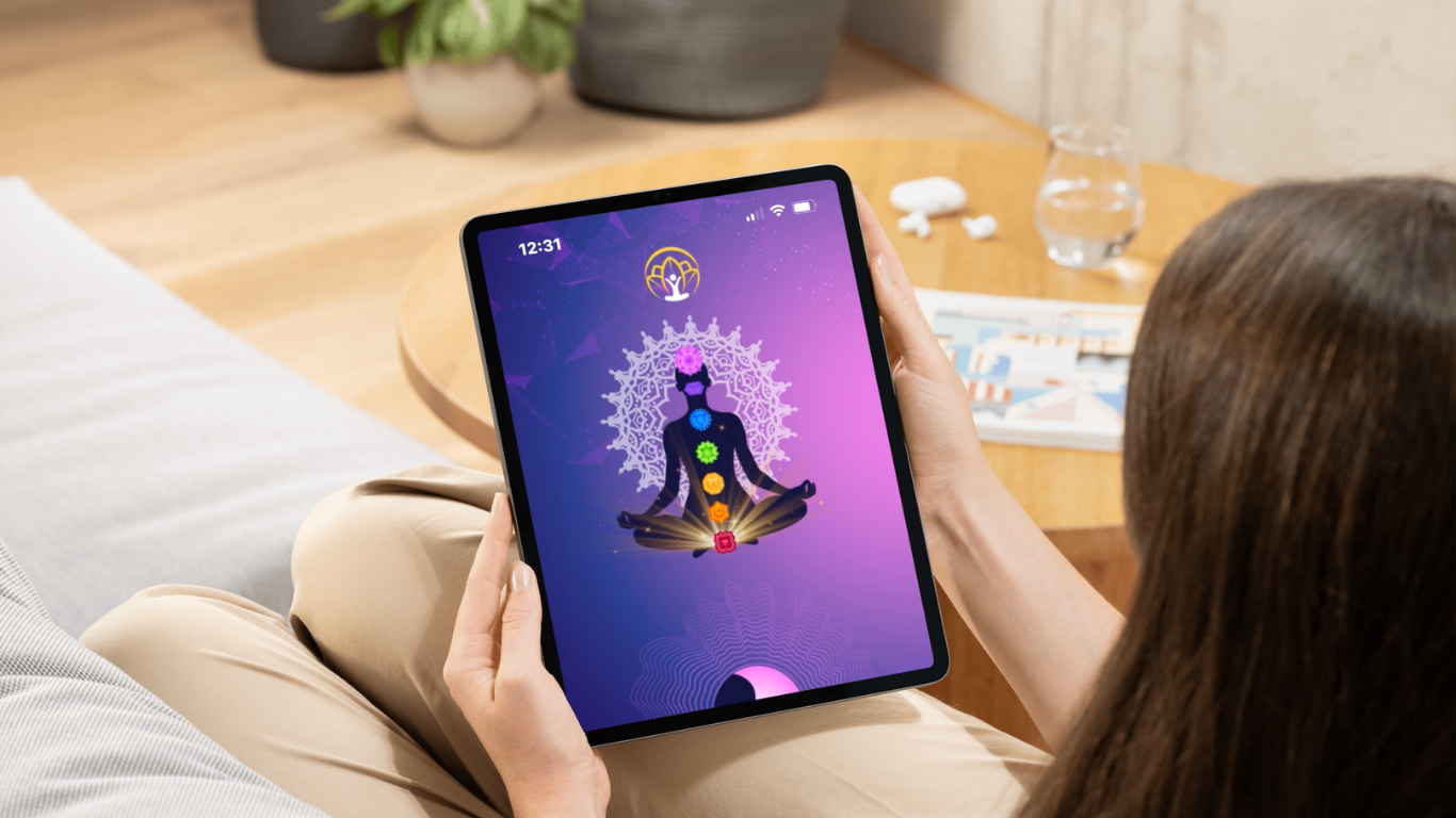 A person holding a tablet with a meditative silhouette and chakra symbols on the screen, sitting on a beige couch in a cozy room.