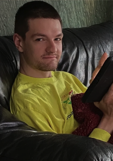 A young man with short dark hair and a light beard, wearing a bright yellow shirt, sitting on a black leather couch and holding a remote control.