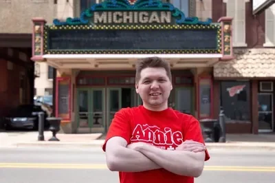 Person standing on a city street in front of a Michigan theater marquee, wearing a red shirt with 'Annie' printed on it, arms crossed, smiling.