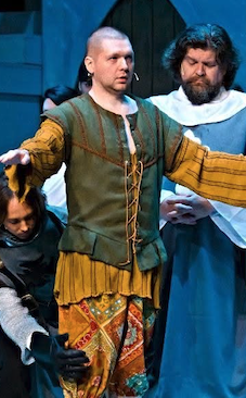 A scene from a theatrical play with three actors in period costumes, one with outstretched arms and a serious expression, and two others observing.