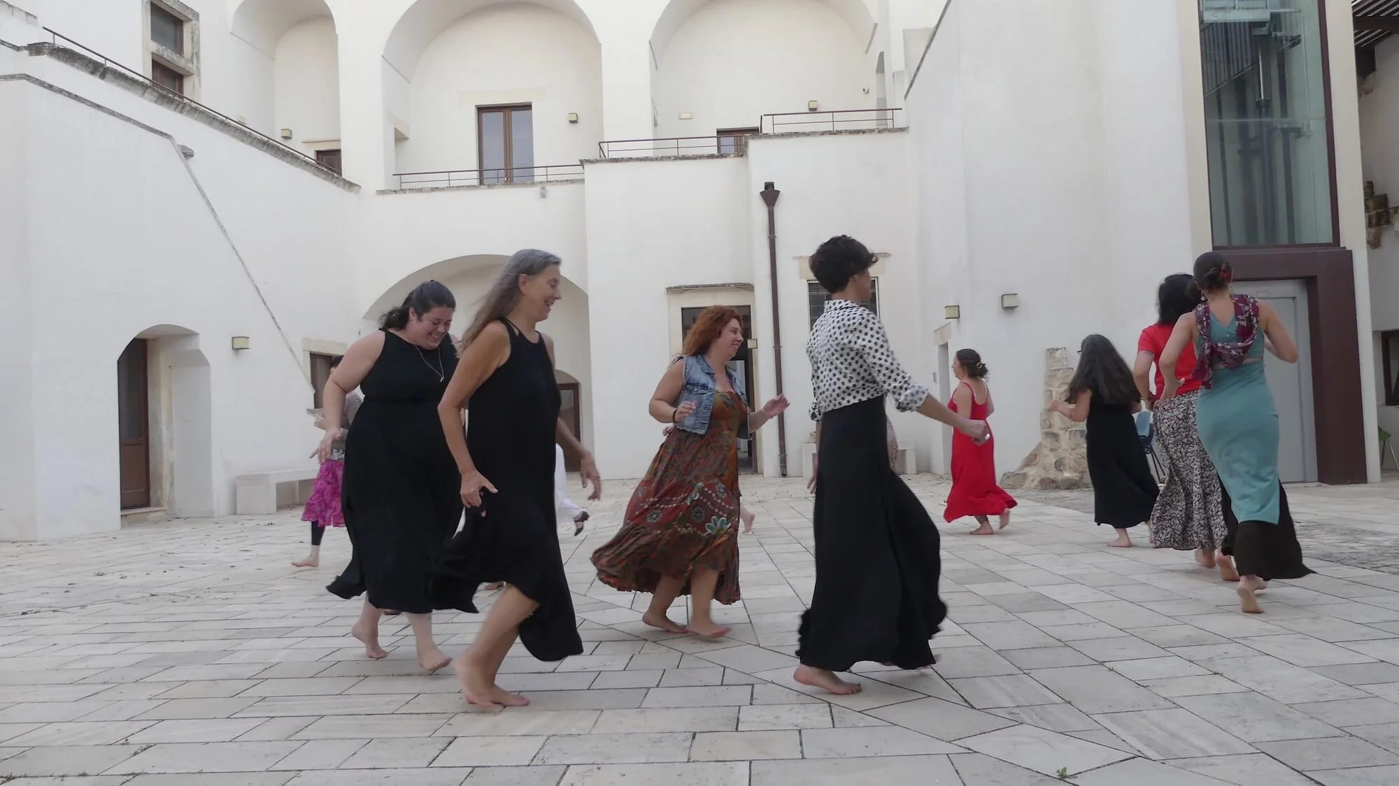Group of women dancing barefoot in an open courtyard with white walls and arched windows. Some women are wearing long skirts and dresses, smiling and enjoying the dance.
