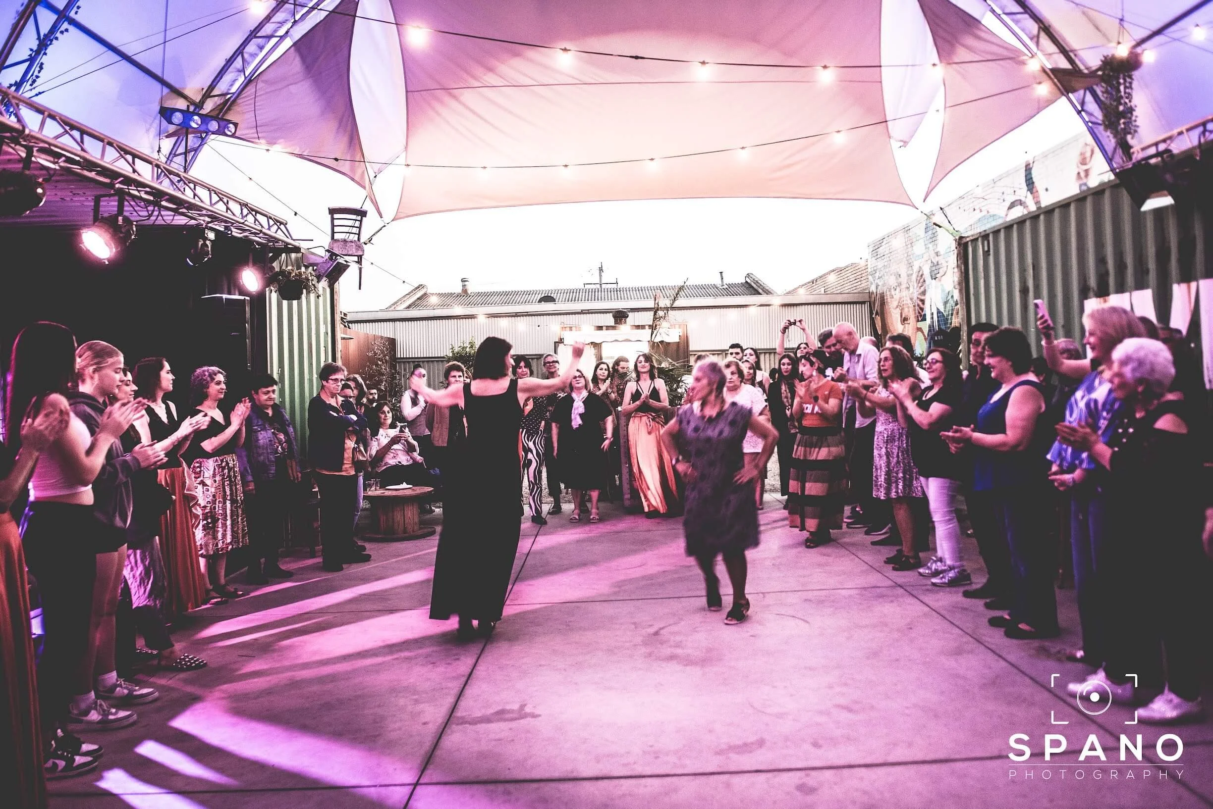 A group of people dancing and socializing at an outdoor celebration under a large canopy with string lights.