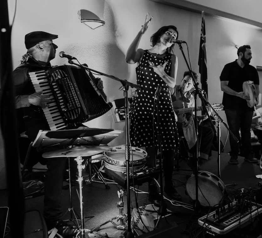 Musicians performing on stage, with a woman singing into a microphone, a man playing an accordion, and others playing percussion instruments in the background.