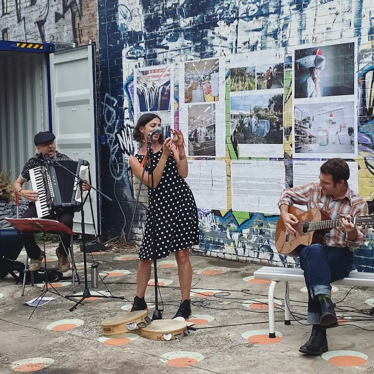 Three street performers playing music outdoors in front of a graffiti-covered wall with photos and posters. One woman is singing with a tambourine on the ground, a man on the left is playing an accordion, and another man on the right is playing an acoustic guitar.