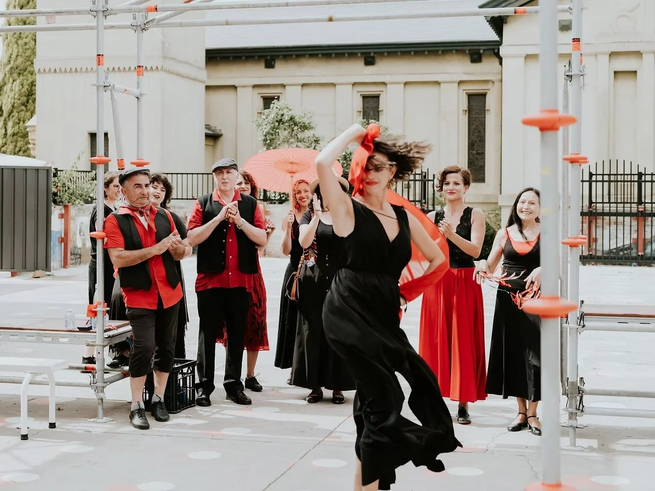 A woman dancing energetically in front of a group of people outside a church, with some clapping and smiling at her performance, using a red scarf and umbrella.