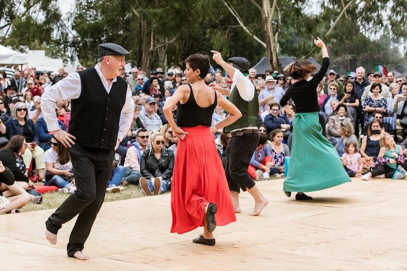 Four people dancing outdoors on a wooden stage with a large crowd watching, some sitting on the grass and others standing, surrounded by trees and tents in the background.